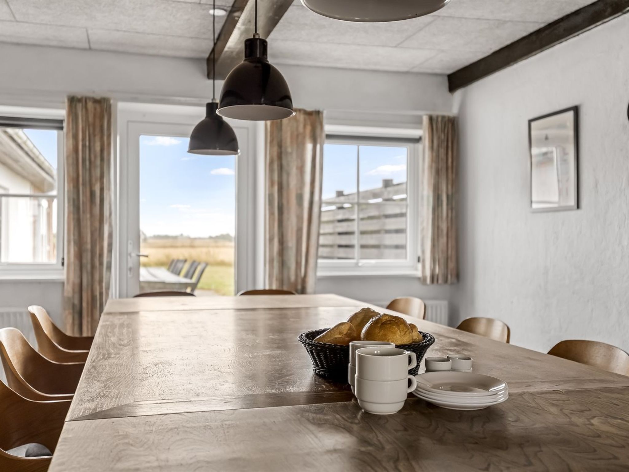 A dining room with a long table, chairs, a hanging light fixture, and a view of the outdoors.