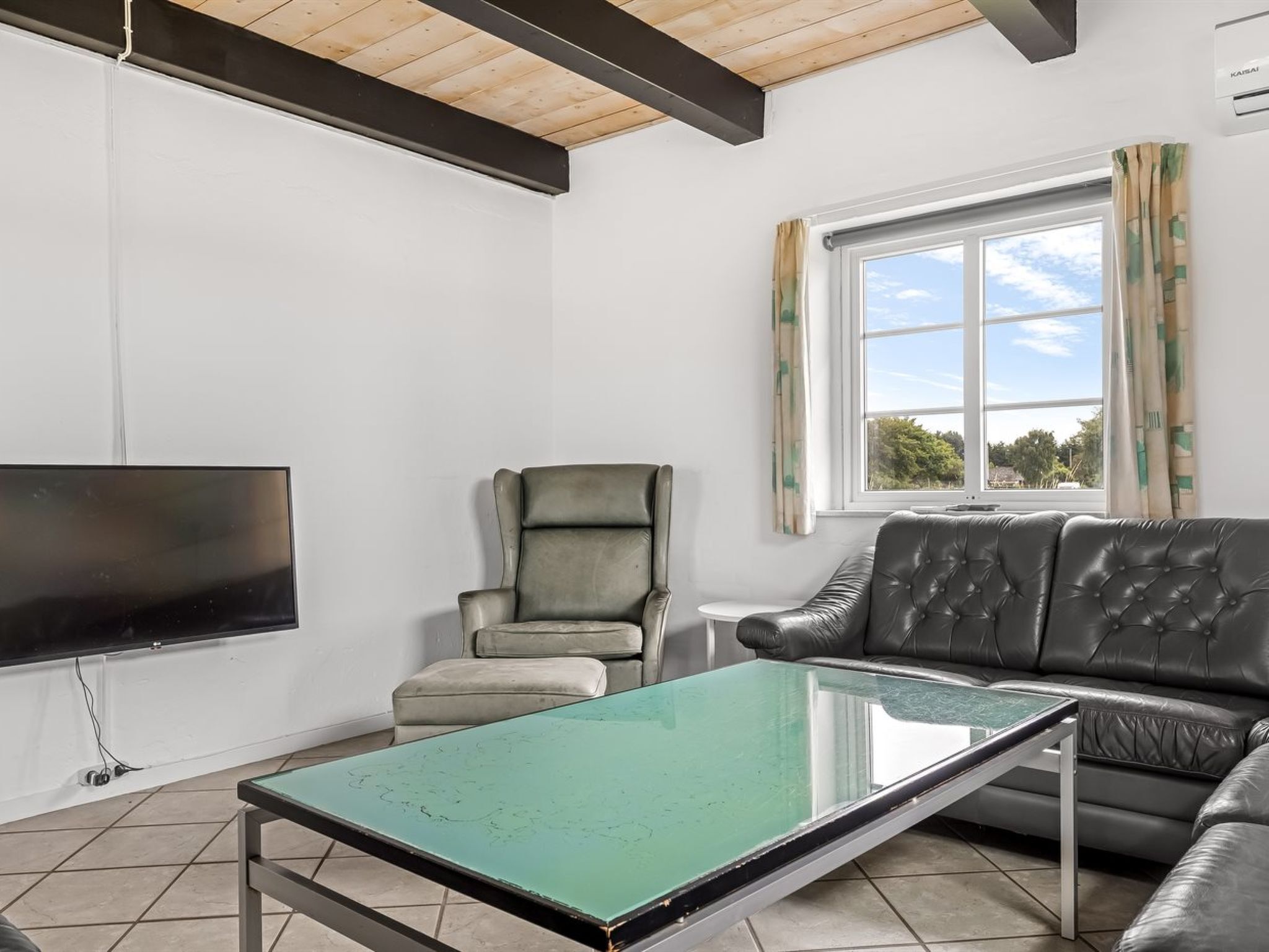 A living room with a flat-screen TV mounted on the wall, a leather sofa, a green coffee table, and a window with curtains.