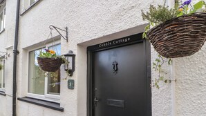 A cottage with a black door, a white textured wall, and two hanging baskets with flowers. - Cobble Cottage (Keswick)