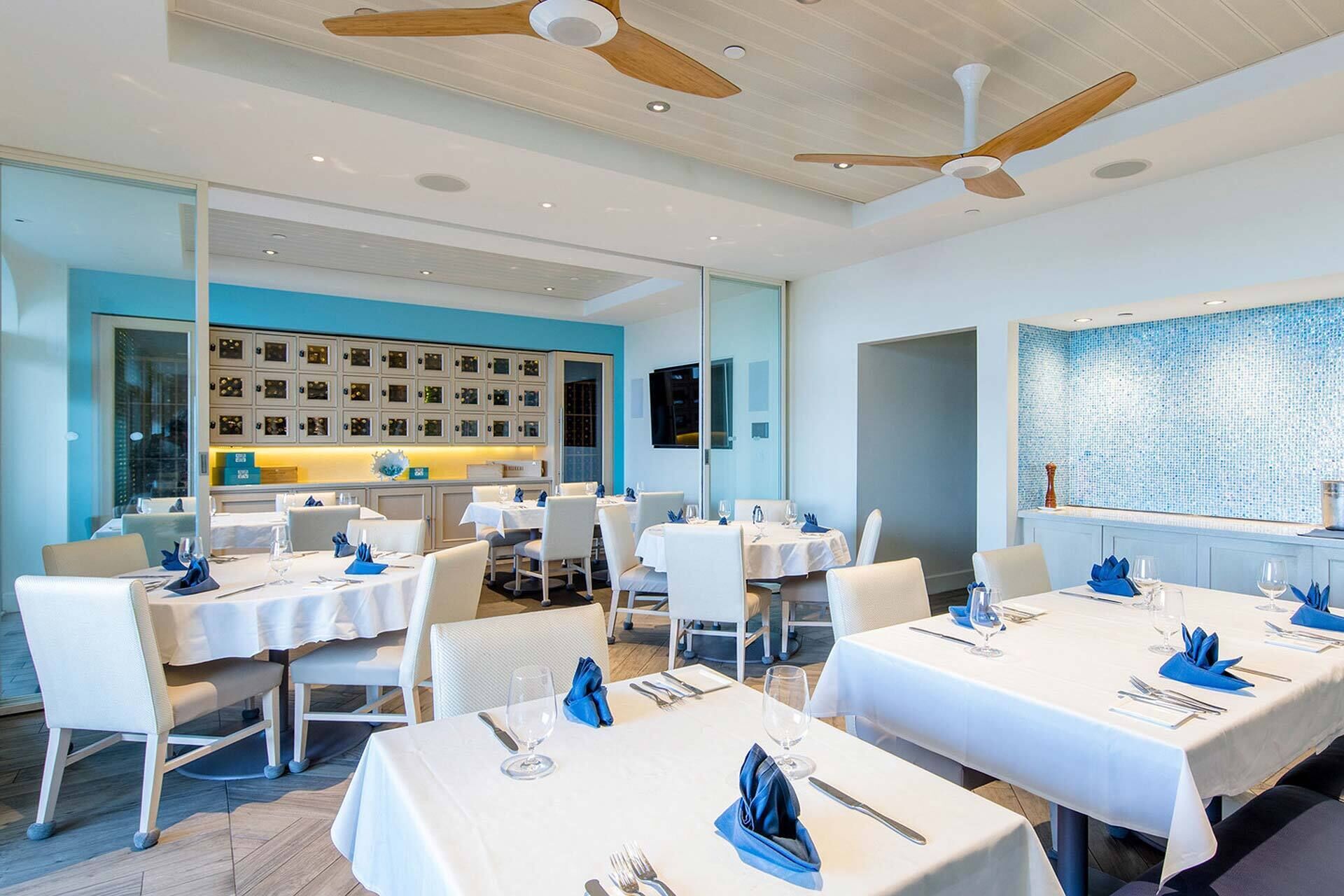 Ein modernes Restaurant mit weißen Tischen und Stühlen, Deckenventilatoren und einer blauen Wand mit gerahmten Bildern.