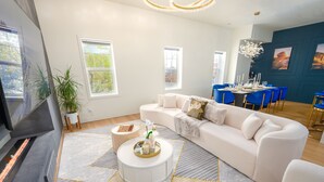 A modern living room with a white sofa, a round coffee table, and a TV. There are windows, a dining area with blue chairs, and a fireplace. - Your luxury vacation begins here! (Drumheller)