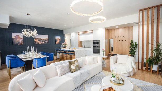 A modern living room with a white sofa, blue chairs, and a chessboard. There is a kitchen in the background with white cabinets and a wooden accent wall. - Your luxury vacation begins here! (Drumheller)
