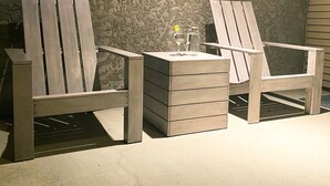 Dos sillas de madera tipo Adirondack con una mesita y un vaso de agua en el centro, ubicadas frente a una pared con textura.
