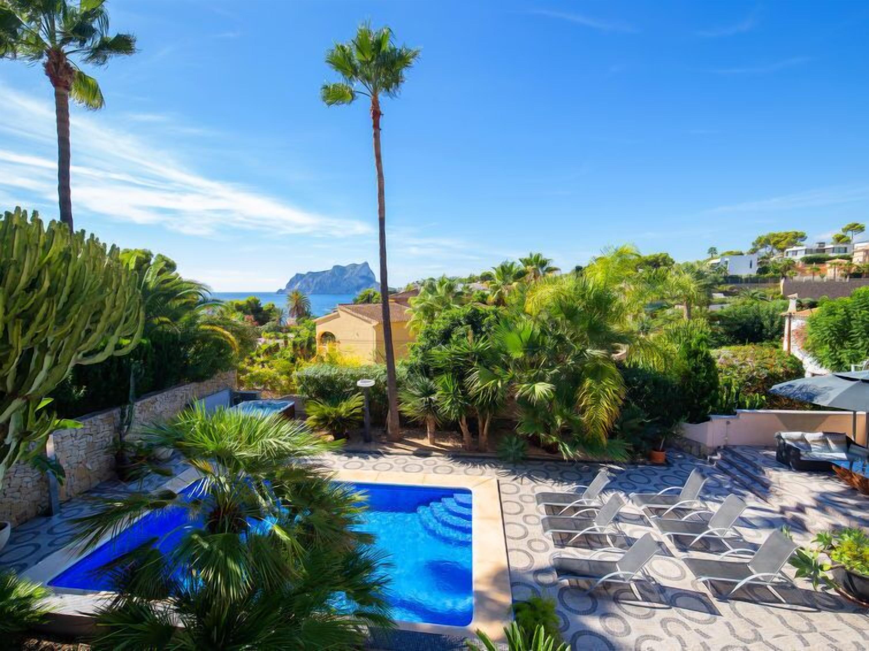 A pool area with lounge chairs, surrounded by palm trees and a view of the sea.