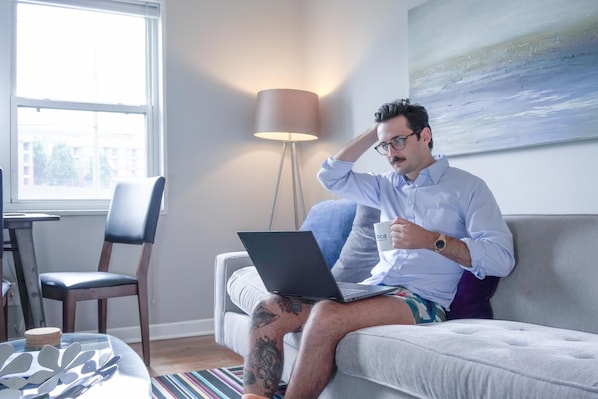 A man sitting on a couch with a laptop, holding a mug, in a room with a lamp, a painting, and a window.