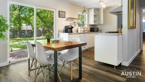 A modern kitchen with white cabinetry, a dining area with a wooden table and white chairs, and a large window overlooking a backyard. - 5412 Pine xxxxxxxxxx (Austin)