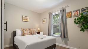 A neatly made bed with a white comforter and colorful pillows, a bedside table with a lamp, and a window with curtains. - 5412 Pine xxxxxxxxxx (Austin)