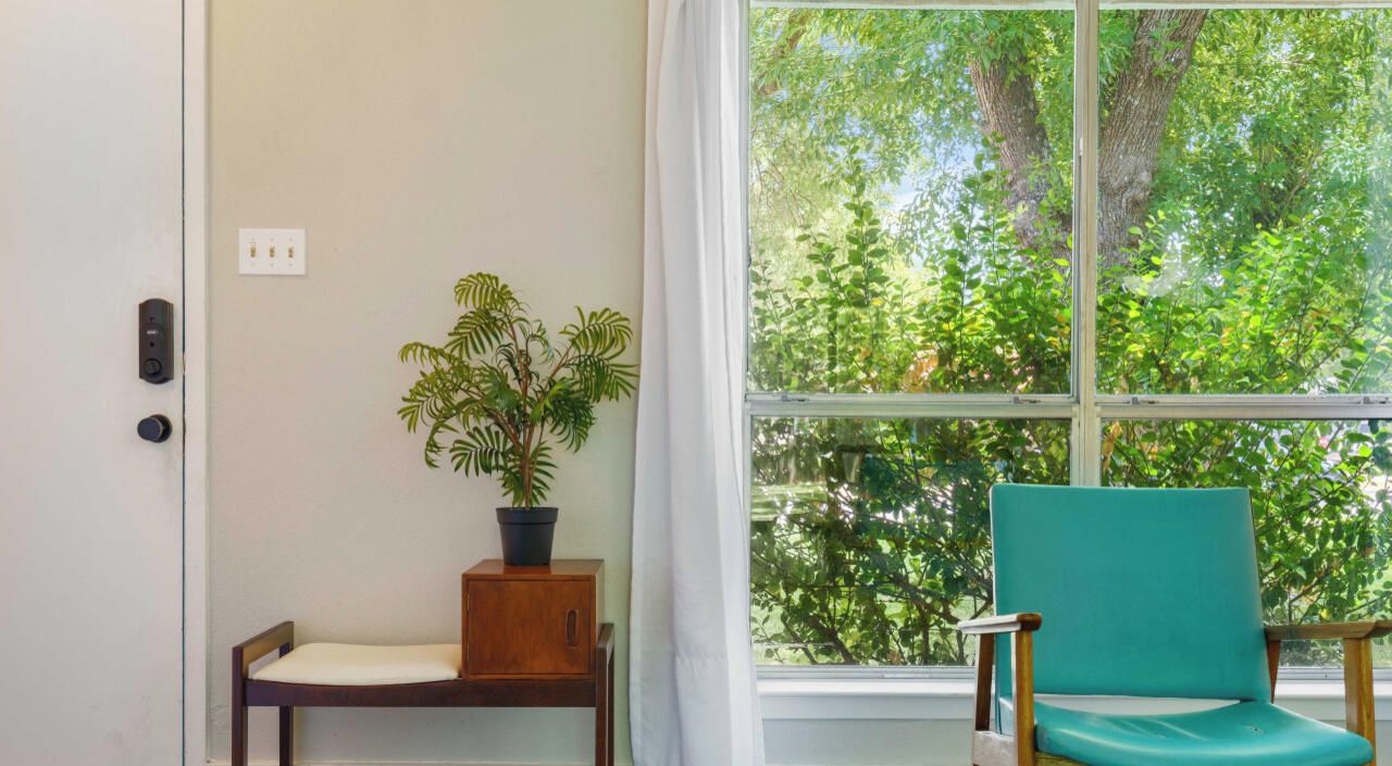 Une pièce avec un banc en bois, une plante en pot et une chaise de couleur turquoise près d’une fenêtre donnant sur des arbres.