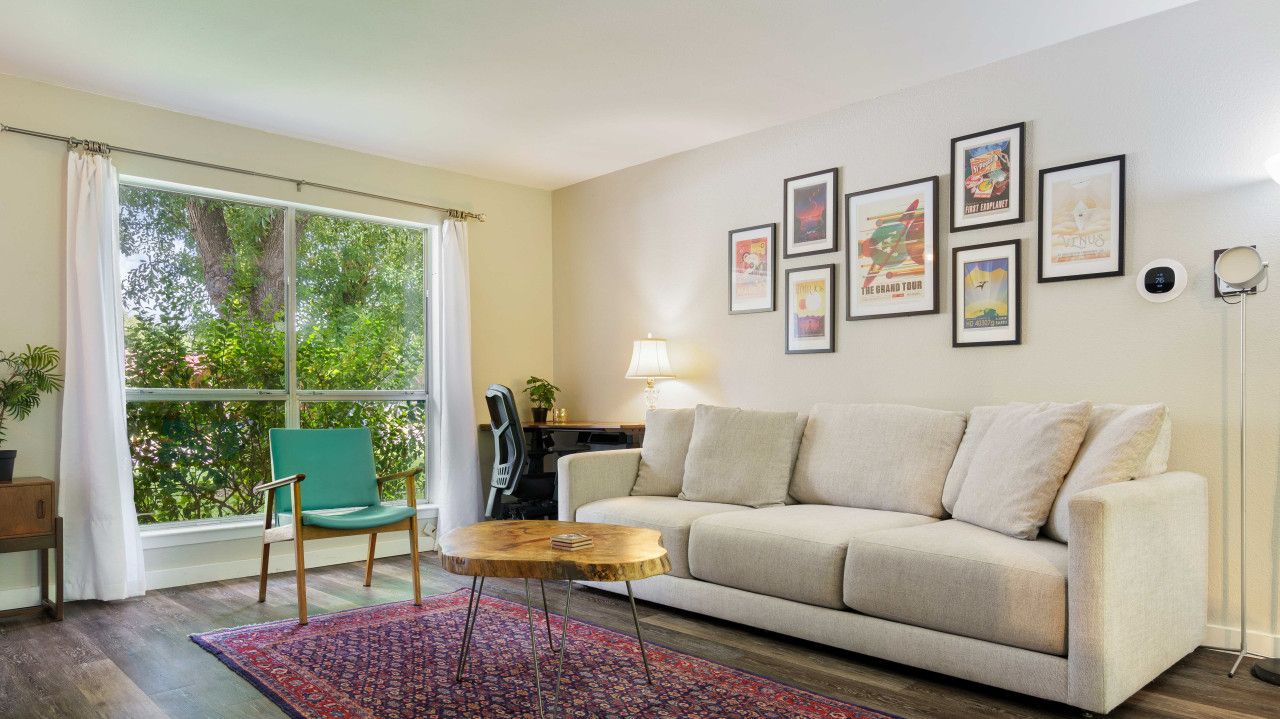 Sala de estar con sofá, una mesa de centro, una silla, alfombra y una ventana con cortinas. Hay cuadros enmarcados en la pared.