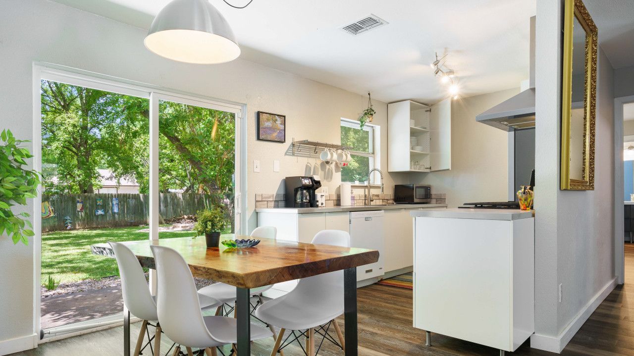 Een moderne keuken met witte kasten, een eethoek met een houten tafel en witte stoelen, en een groot raam met uitzicht op een achtertuin.