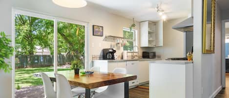Una cocina moderna con armarios blancos, un comedor con una mesa de madera y sillas blancas, y un amplio ventanal con vistas a un jardín trasero.