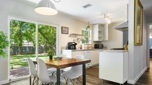 Una cocina moderna con armarios blancos, un comedor con una mesa de madera y sillas blancas, y un amplio ventanal con vistas a un jardín trasero.
