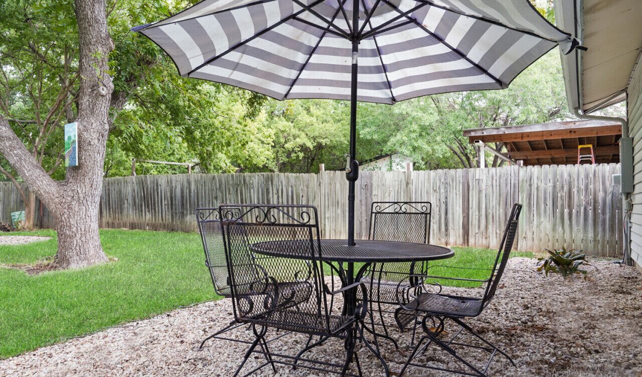 Une terrasse avec une table et des chaises en métal, un parasol à rayures et, au fond, une clôture en bois.
