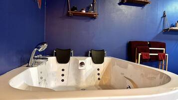 Una bañera grande con dos sillas negras, un lavamanos blanco y una pared azul con estantes que exhiben diversos objetos.