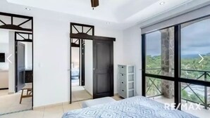 Une chambre dotée d’un grand lit, d’une armoire, d’un bureau et offrant une vue sur la verdure grâce à de grandes fenêtres.