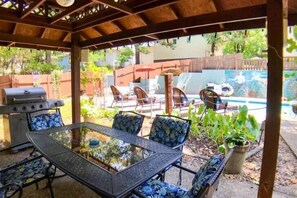 An outdoor patio with a glass-top table and chairs, a grill, and a pool in the background. - The Grand Suite at Tradewinds (Eureka Springs)