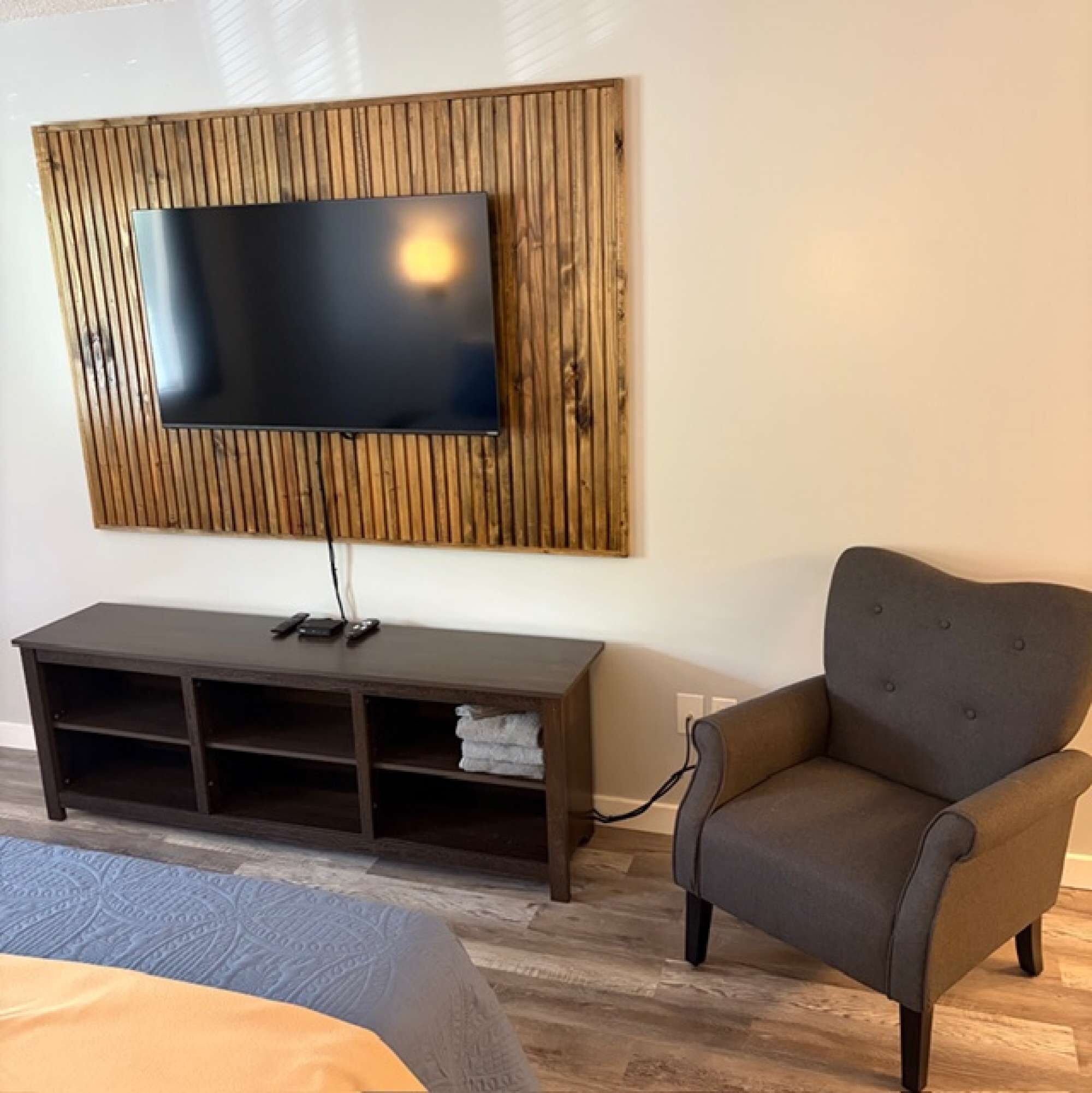 A room with a flat-screen TV mounted on a wooden panel, a dark brown entertainment unit, a grey armchair, and a bed with patterned bedding.