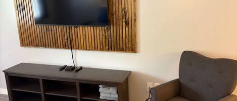 A room with a flat-screen TV mounted on a wooden panel, a dark brown entertainment unit, a grey armchair, and a bed with patterned bedding.