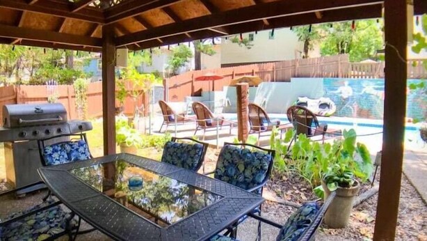 Een terras buiten met een tafel en stoelen met een glazen blad, een barbecue en een zwembad op de achtergrond.