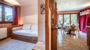 Ein Zimmer mit einem Bett, einem Esstisch mit Stühlen, einem Fenster mit Vorhängen und Blick ins Freie.