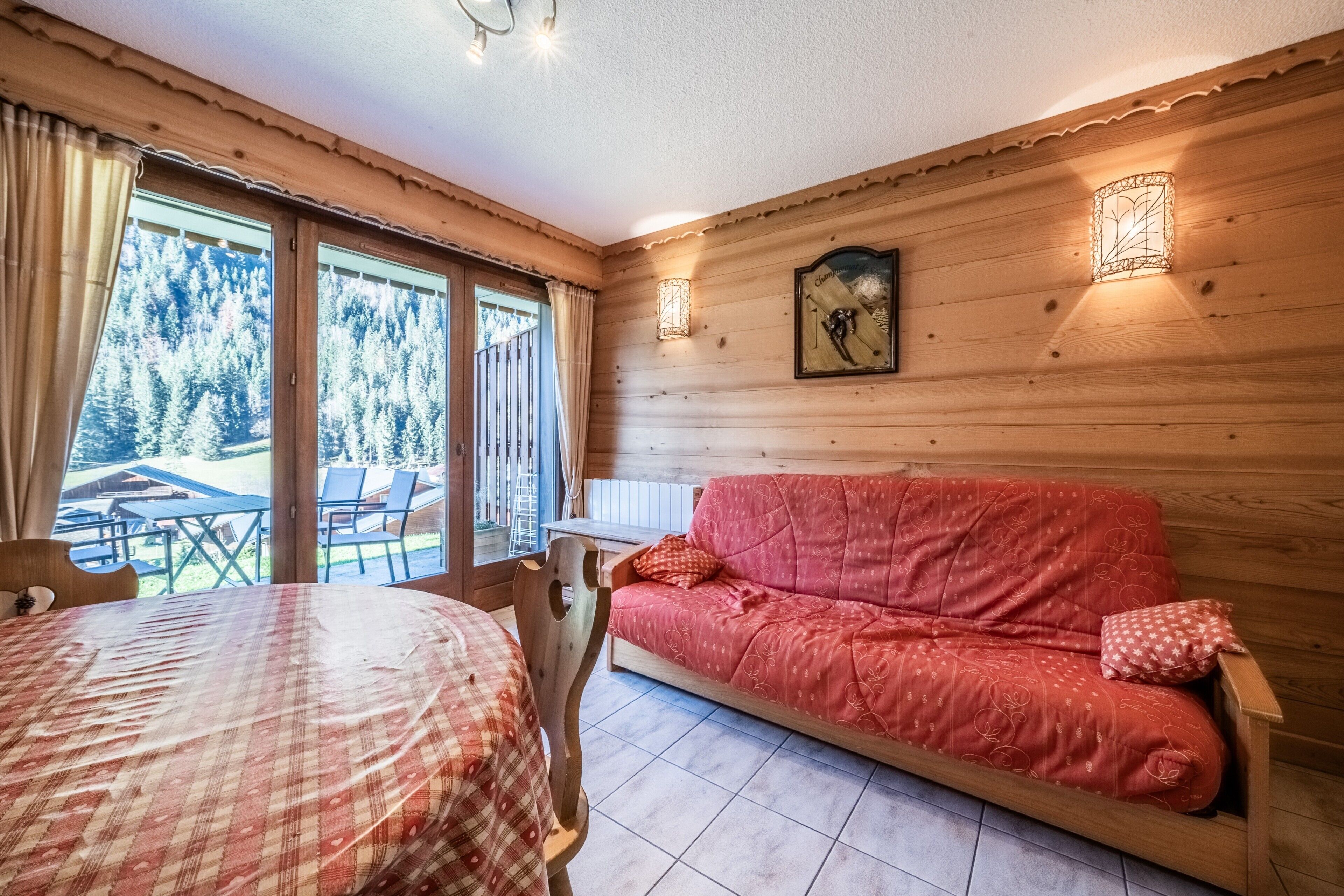 L’intérieur d’un chalet en bois, avec un canapé rouge, une table à manger et une vue sur une forêt, visible à travers de grandes fenêtres.