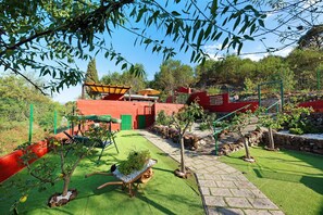 Un jardin avec un petit arbre, une brouette remplie de plantes et un bâtiment rouge.