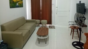 A living room with a beige sofa, two matching ottomans, a wooden coffee table, a brown curtain, a white door, a black fan, and a painting on the wall. - Omah Bondi Family Homestay by FH Stay (Yogyakarta)