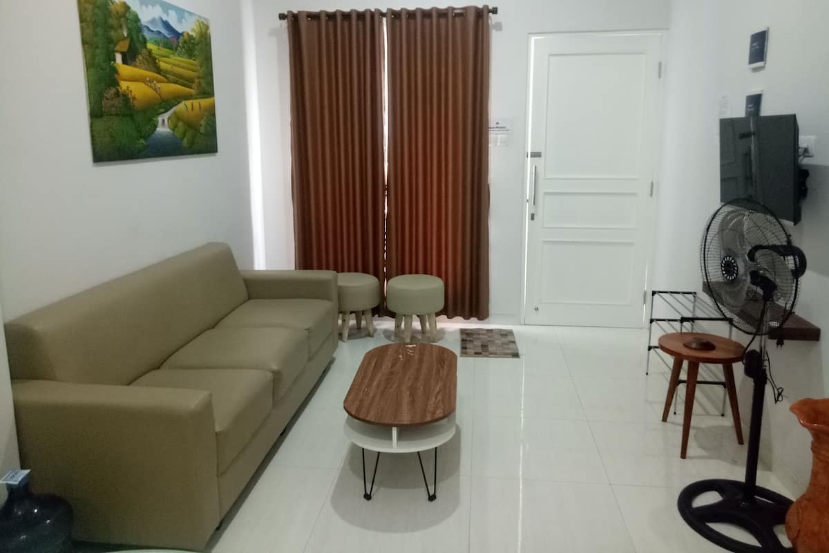 A living room with a beige sofa, two matching ottomans, a wooden coffee table, a brown curtain, a white door, a black fan, and a painting on the wall.