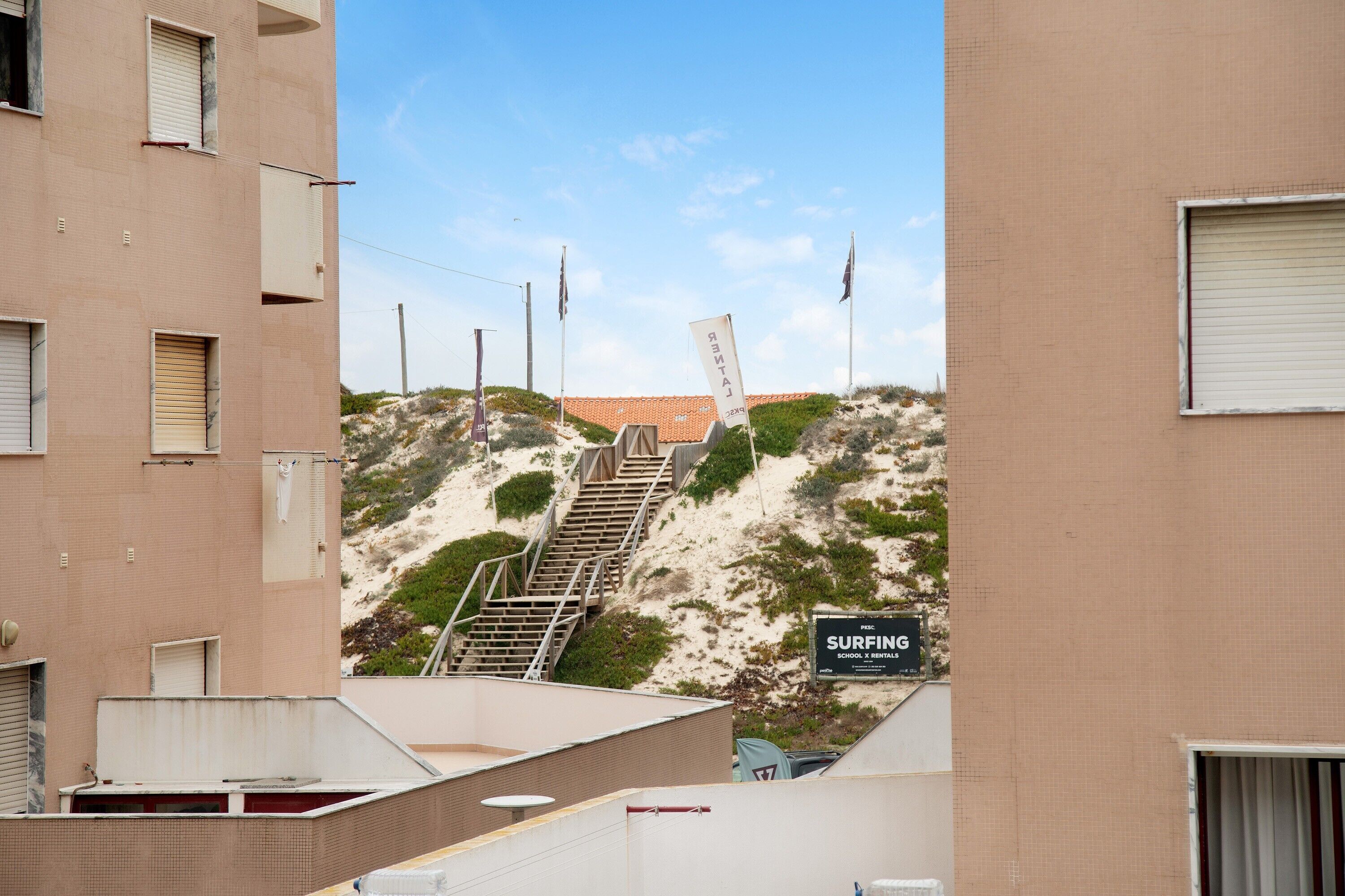 Ein Gebäude mit einem Schild für eine Surfschule, einer Treppe zu einem Sandbereich und einem klaren Himmel.