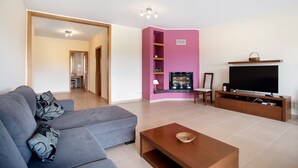 Een woonkamer met een grijze bank, een houten salontafel, een flatscreen televisie op een houten dressoir, en een roze accentmuur.