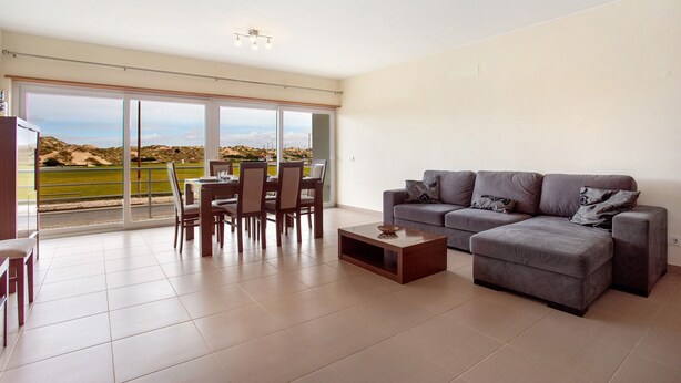 Een ruime woonkamer met een grote schuifpui naar een balkon, een grijze bank en een eettafel met stoelen.