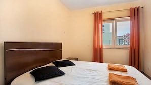 Een hotelkamer met een bed, twee kussens, een raam met gordijnen en uitzicht op gebouwen.