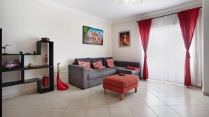 Un salon avec un canapé gris, des rideaux rouges, un pouf rouge, une table basse en verre et un tableau accroché au mur.