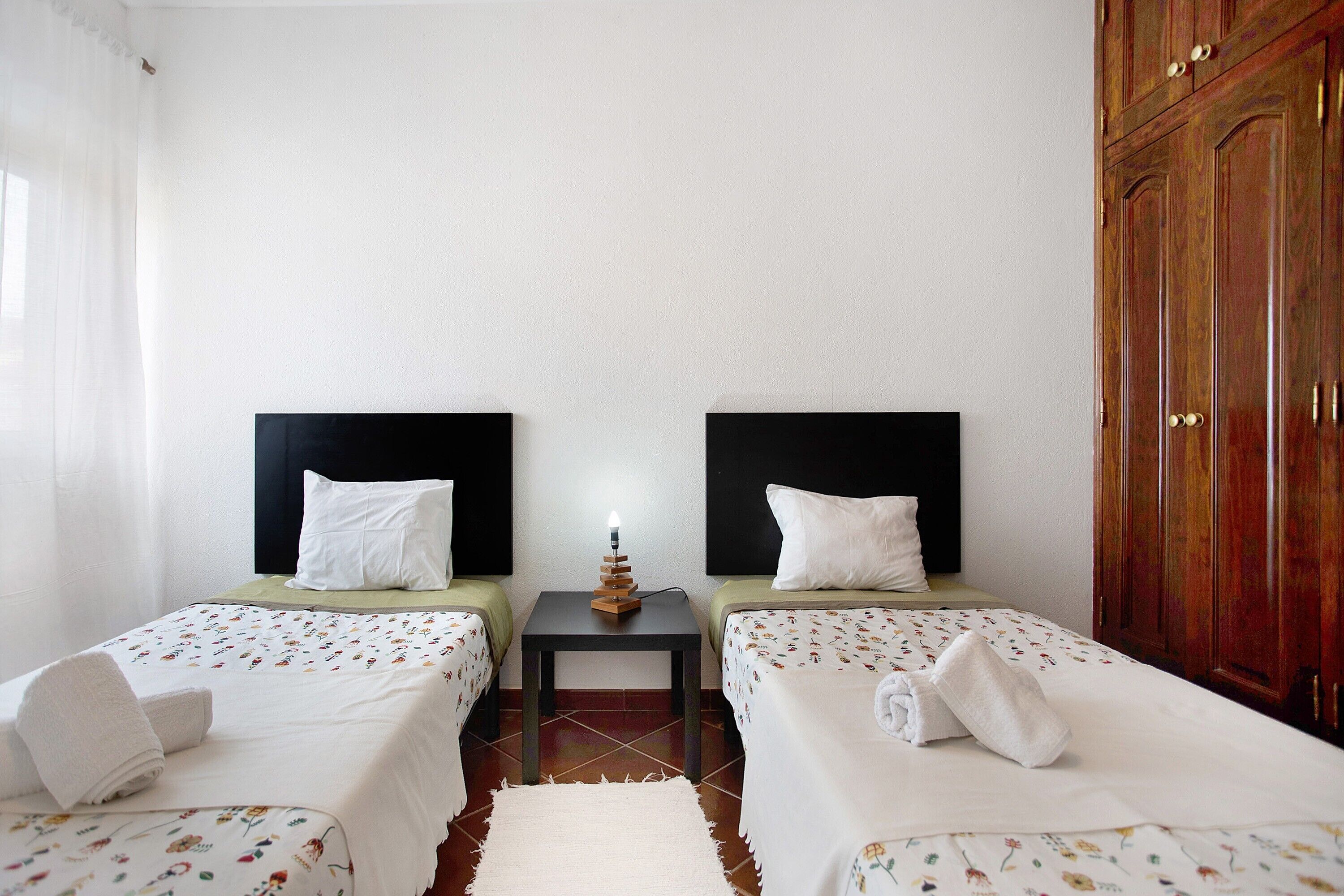 Habitación de hotel con dos camas individuales, una mesita de noche con lámpara, un armario de madera y una cortina blanca.