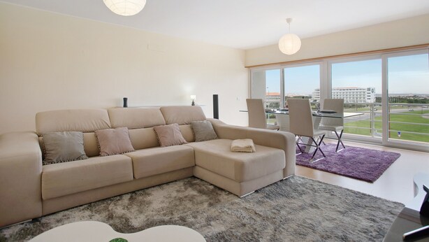 Een moderne woonkamer met een beige hoekbank, een glazen salontafel en een groot raam met uitzicht op een gebouw en een weg.