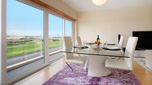 Een moderne eetruimte met een glazen tafel, witte stoelen en uitzicht op een weg en groen.