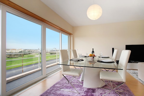Un espace de salle à manger moderne avec une table en verre, des chaises blanches et une vue sur une route et la verdure.
