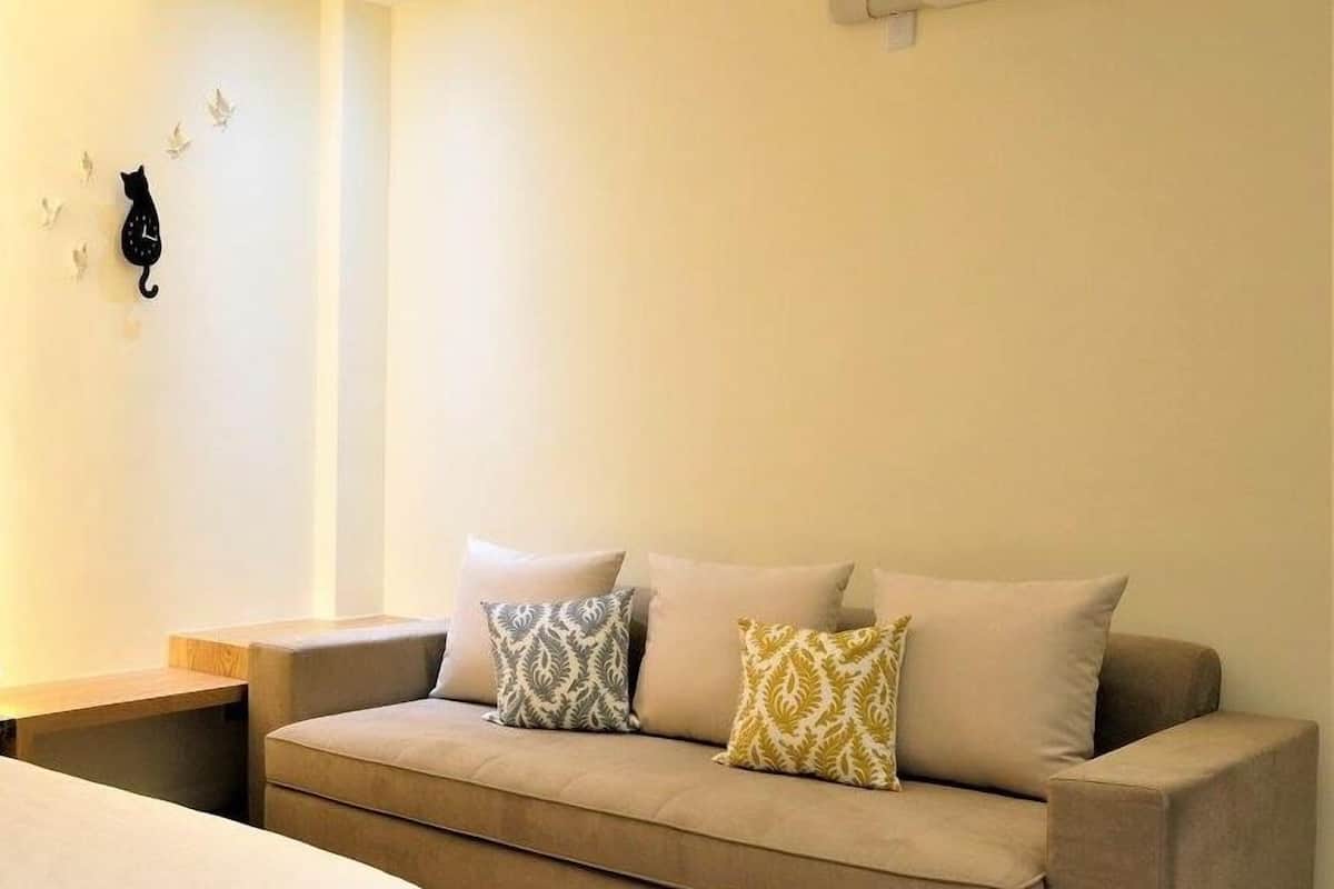 A neutral-colored sofa with patterned cushions, a wooden bedside table, and a white bed in a well-lit room with an air conditioning unit on the wall.