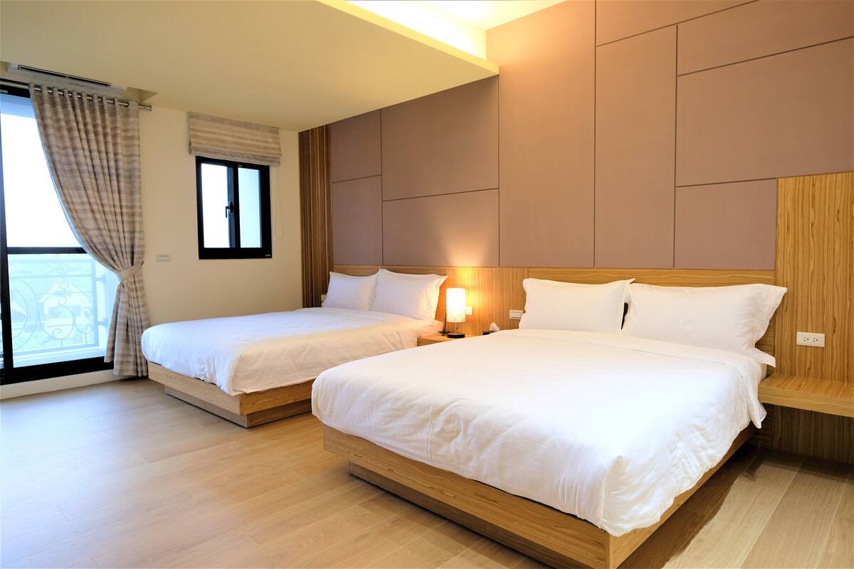 A hotel room with two beds, a large window with curtains, and a wooden headboard.