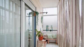 A balcony with a sliding glass door, a potted plant, a small wooden stool, a bench with cushions, and a view of greenery through the window. - Apartment 'Casa Da Mãe Maria' with Sea View, Shared Terrace and Wi-Fi (Buarcos)