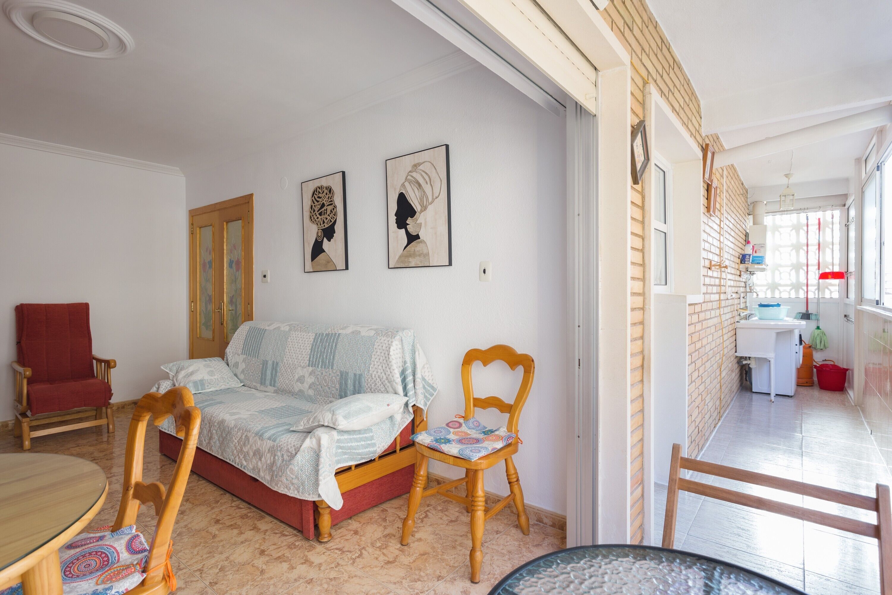 A room with a bed, a wooden chair, a dining table, and a kitchen area visible through an open door.