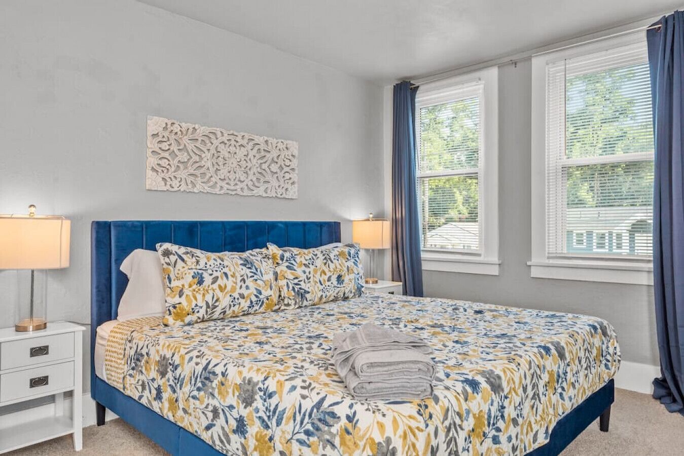Une chambre à coucher avec une tête de lit bleue, recouverte de tissu, une couvre-lit à motifs floraux, deux lampes de chevet, une table de chevet blanche et une fenêtre avec des rideaux bleus.