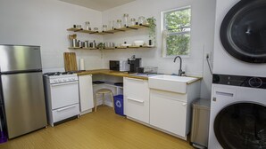 Cocina compacta con refrigerador de acero inoxidable, estufa a gas, microondas y lavadora integrada. Hay repisas de madera sobre la encimera y una ventana que proporciona luz natural.