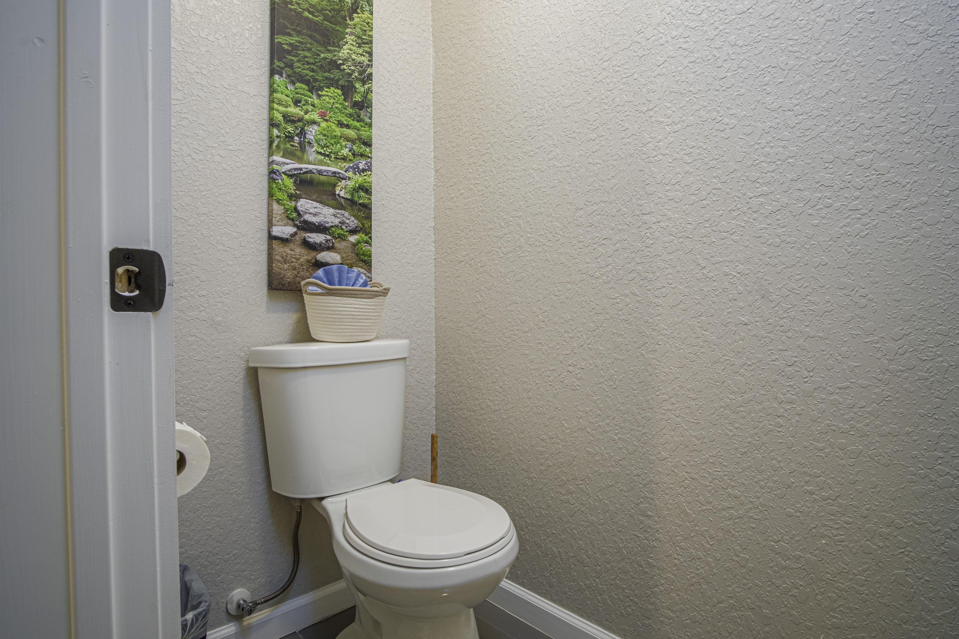 A bathroom with a toilet, a wall-mounted trash can, and a framed picture of a garden.