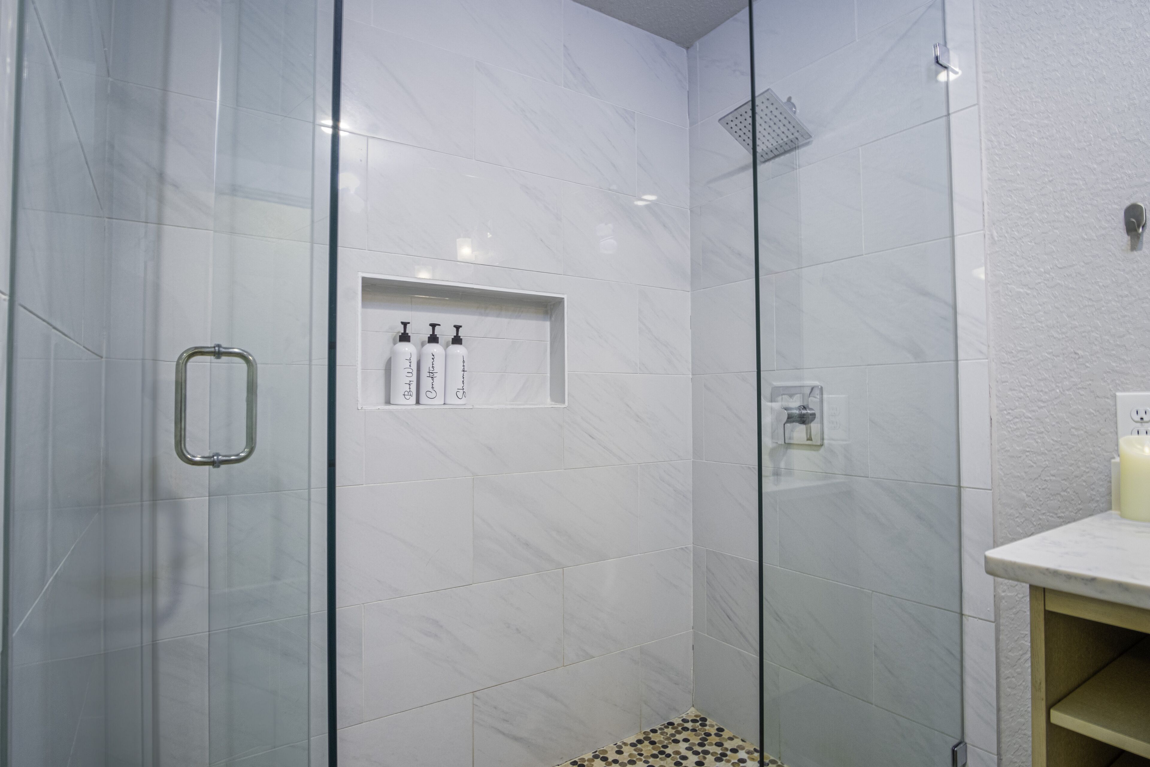 A modern bathroom with white marble tiles, a glass shower enclosure, and a shelf with toiletries.