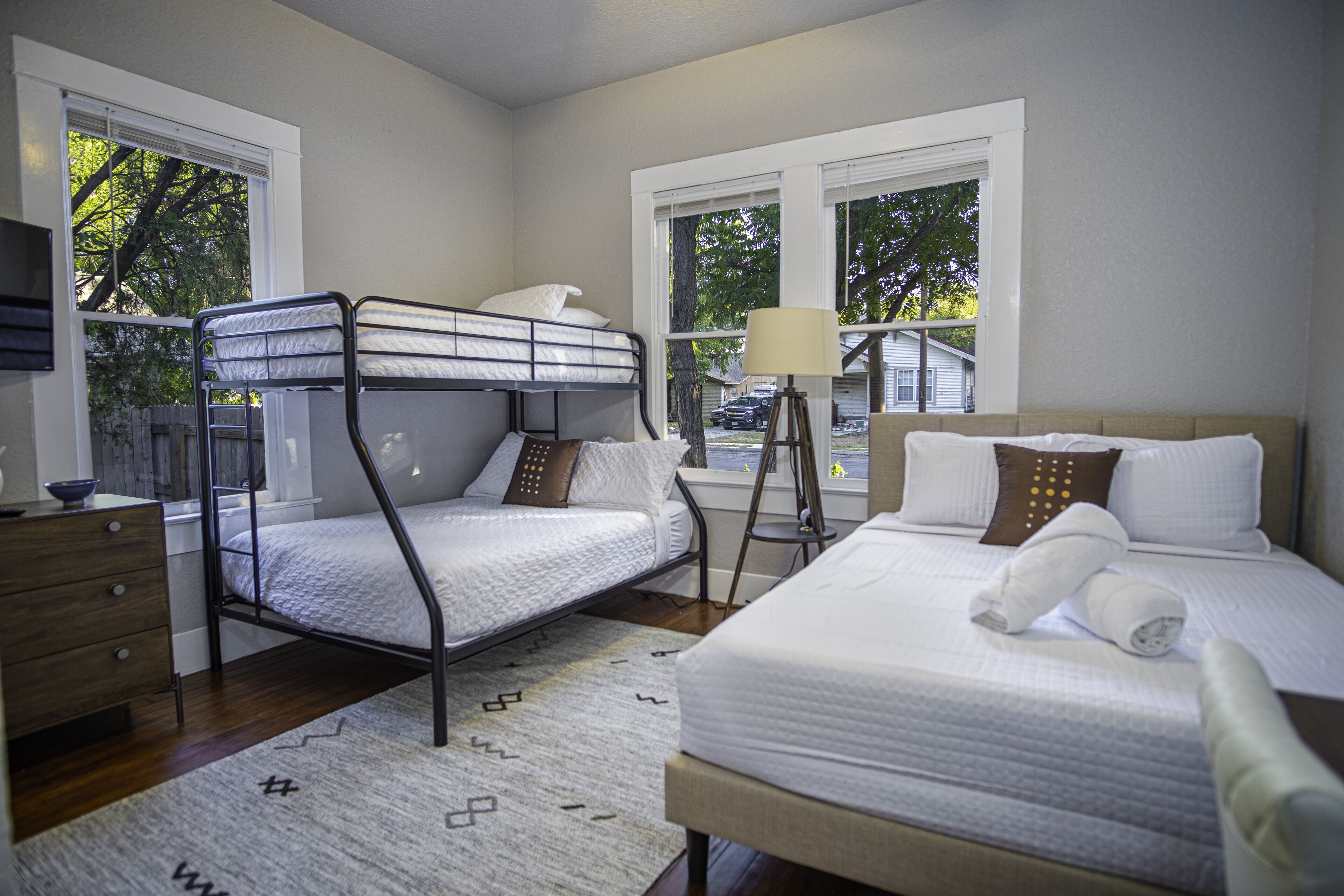 A bedroom with a bunk bed, a single bed, a bedside table, a lamp, and a view of the outside.