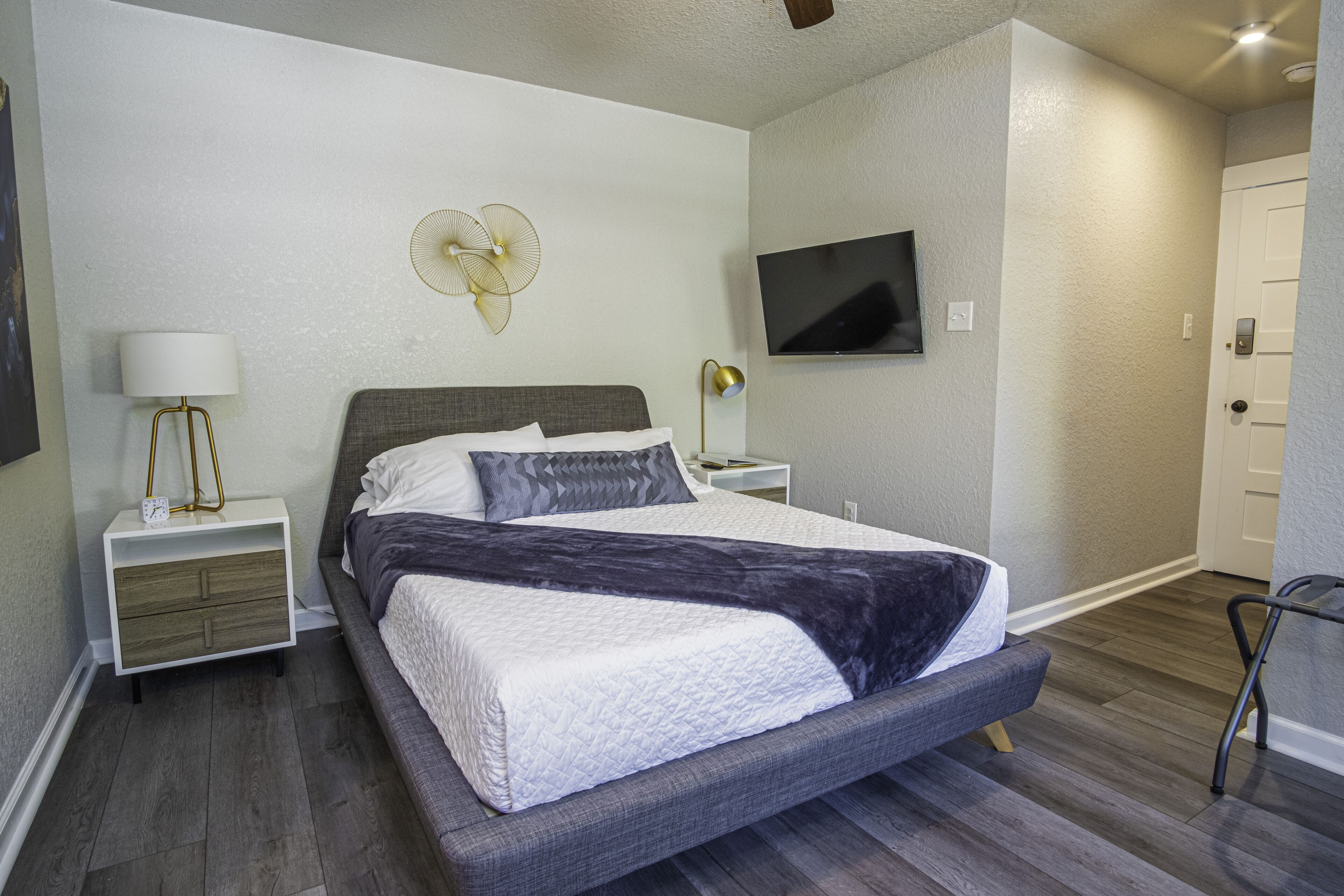 A modern bedroom with a large bed, a bedside table with a lamp, a wall-mounted TV, and a door.