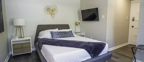 A modern bedroom with a large bed, a bedside table with a lamp, a wall-mounted TV, and a door.