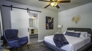 Um quarto com cama, uma cadeira, ventilador de teto, luminária na parede e um quadro.
