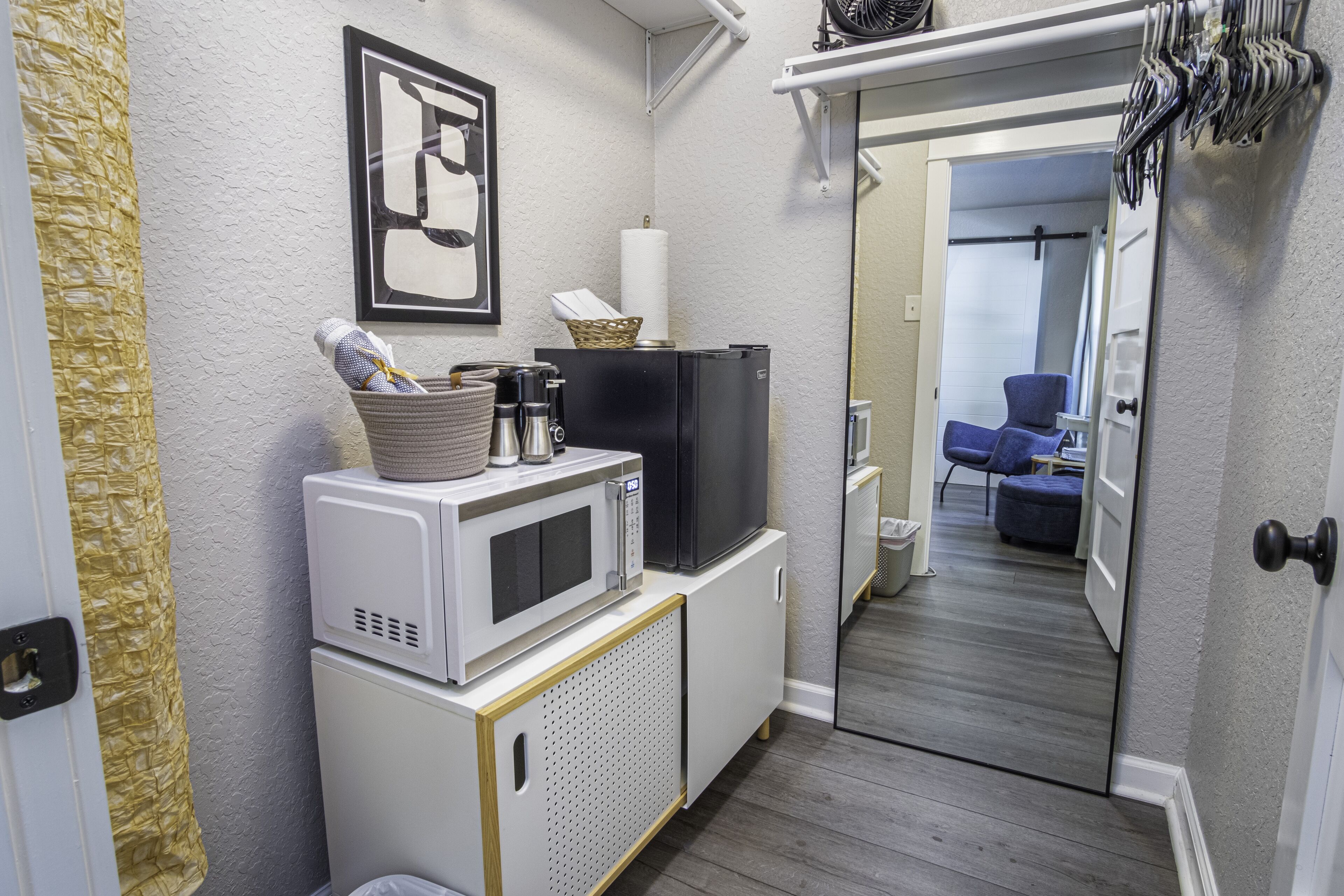 A compact kitchen with a microwave, refrigerator, and a small cabinet. A basket with a towel and a bottle is on the microwave. A framed picture hangs on the wall.